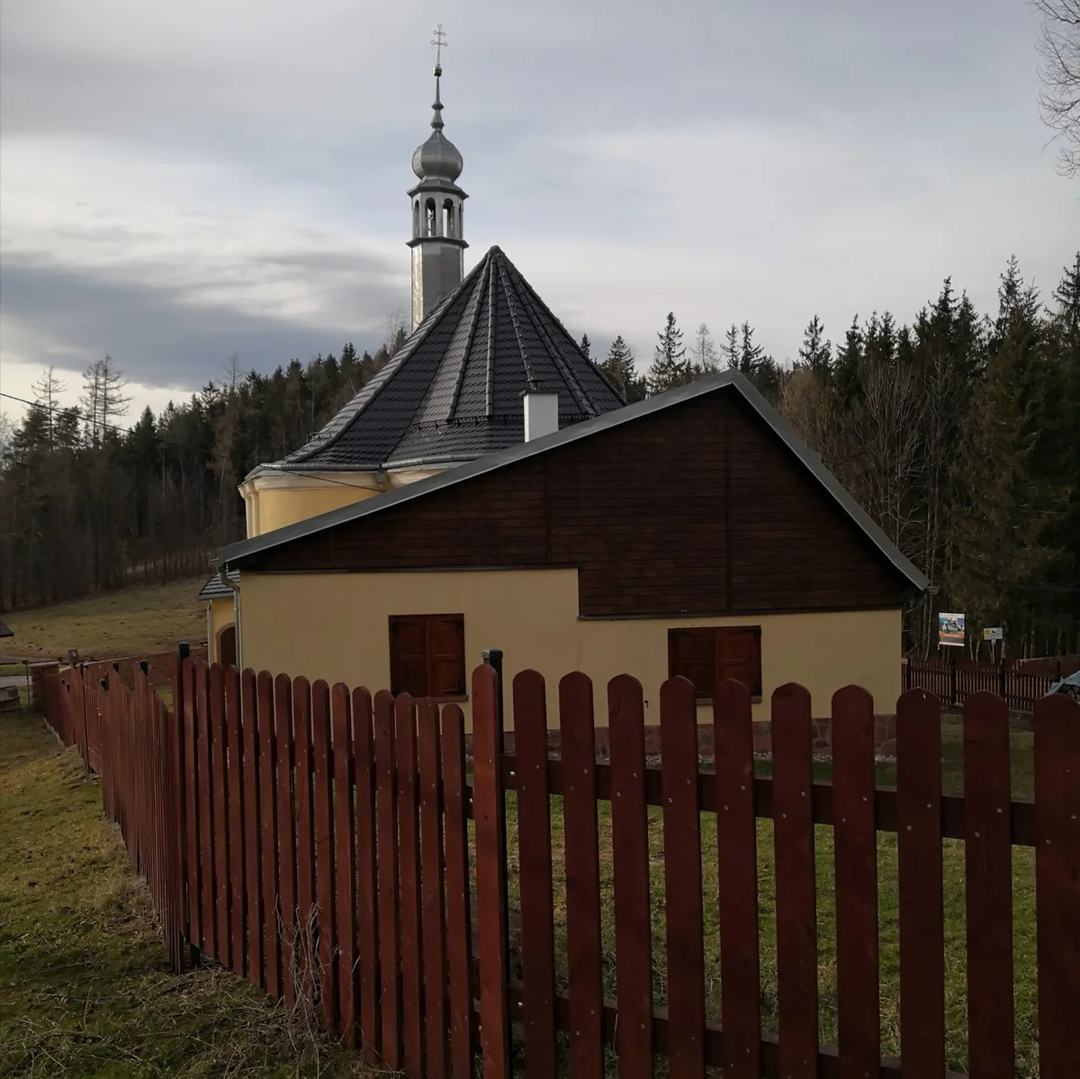 Budynek o tradycyjnym kolorze widoczny zza płotu letnią porą.